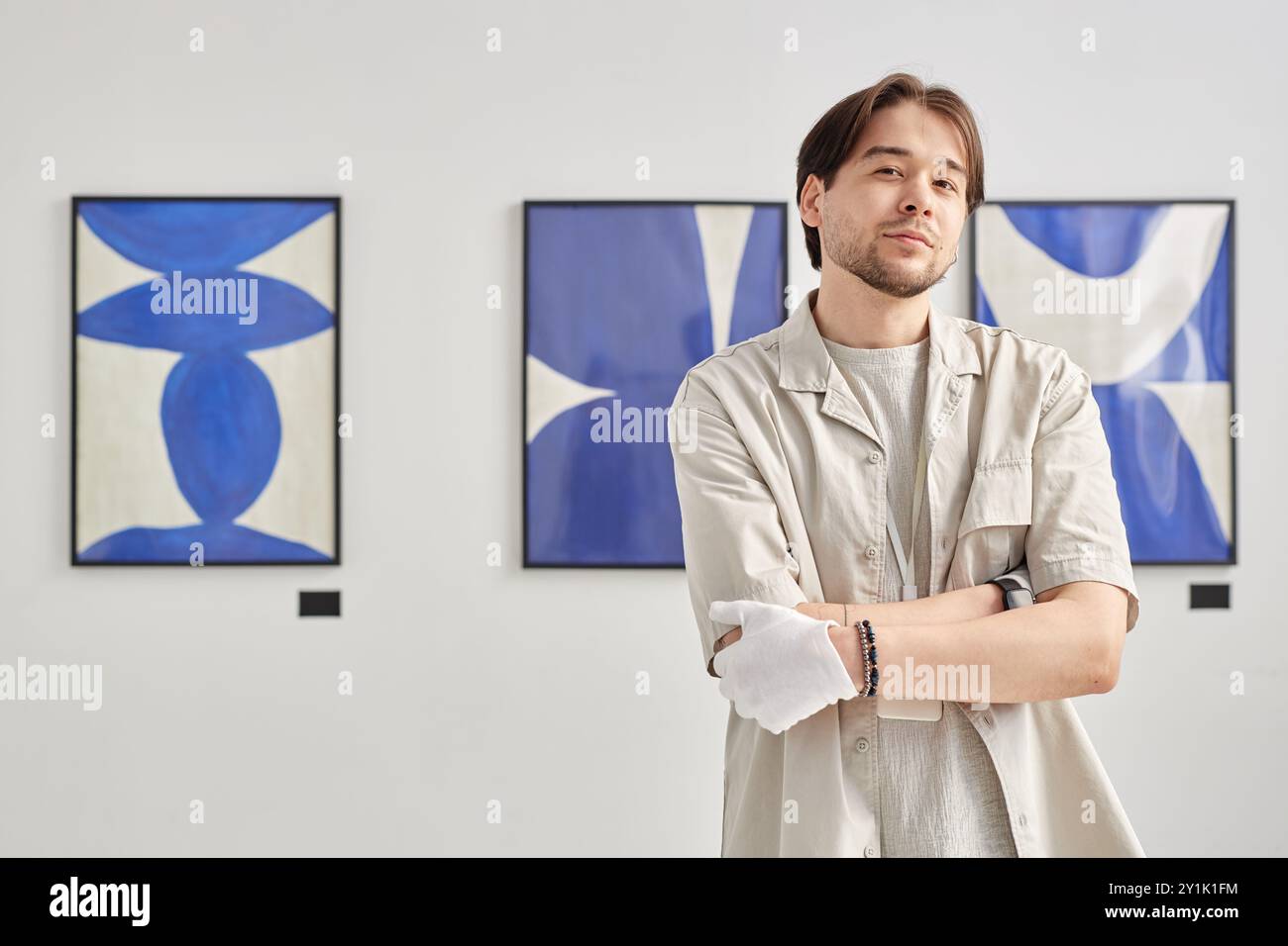 Waist up portrait of young man as art gallery curator looking at camera and wearing white gloves while standing with arms crossed against abstract paintings in background copy space Stock Photo