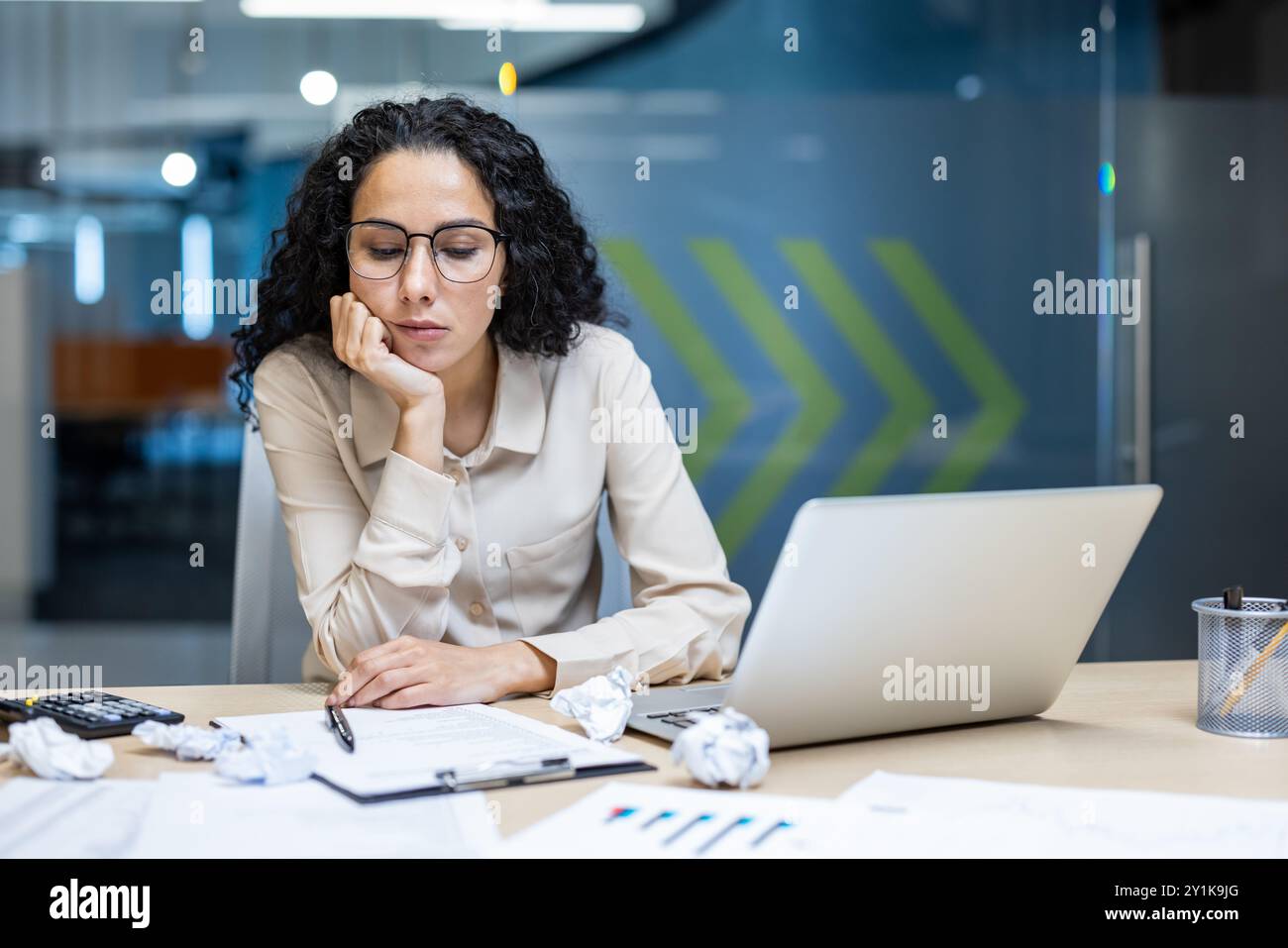 Hispanic businesswoman looking exhausted working on laptop in office. Surrounded by crumpled papers and documents, expressing stress and fatigue. Concept of overwork, deadline pressure Stock Photo