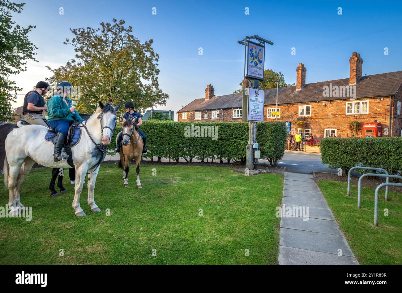 Horse riders enjoy a drink in the saddle at the Hatton Arms pub in rural Stretton near Warrington bathed in evening sunshine. Stock Photo