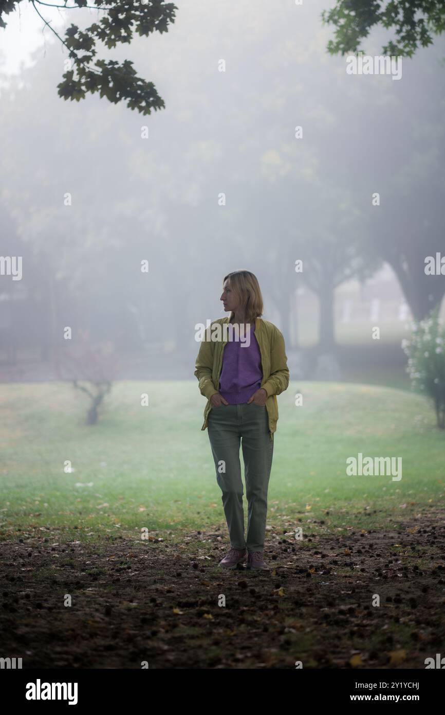 A woman in jeans and a lilac sweater stands in a misty city park, surrounded by fog and soft light, creating a serene and peaceful atmosphere. Stock Photo