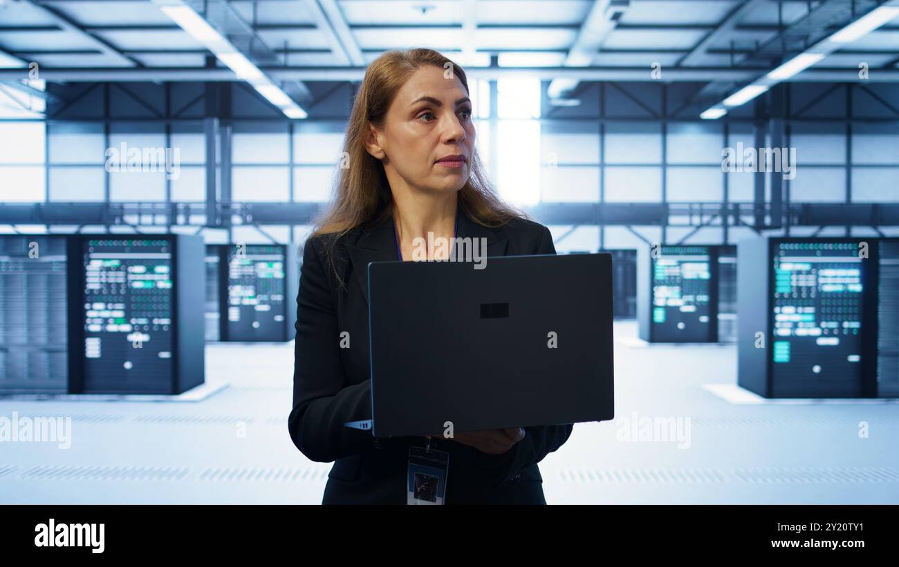 Software developer walking in data center, overseeing server operations using laptop. Woman ensuring optimal performance and data integrity in server farm using notebook, camera B Stock Photo