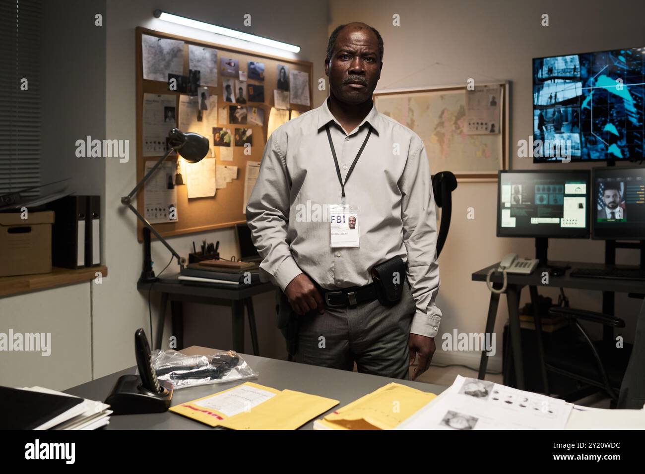 Portrait of African American FBI agent wearing badge, standing in a dimly lit office with various screens and bulletin board in background examining documents Stock Photo