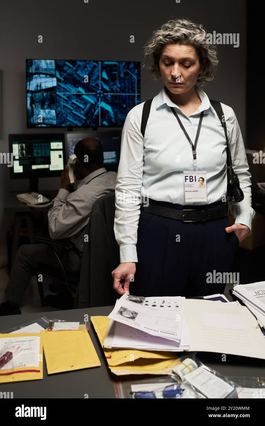 Gray-haired woman with FBI badge reviewing documents at office desk, team member analyzing data on monitors in background Stock Photo