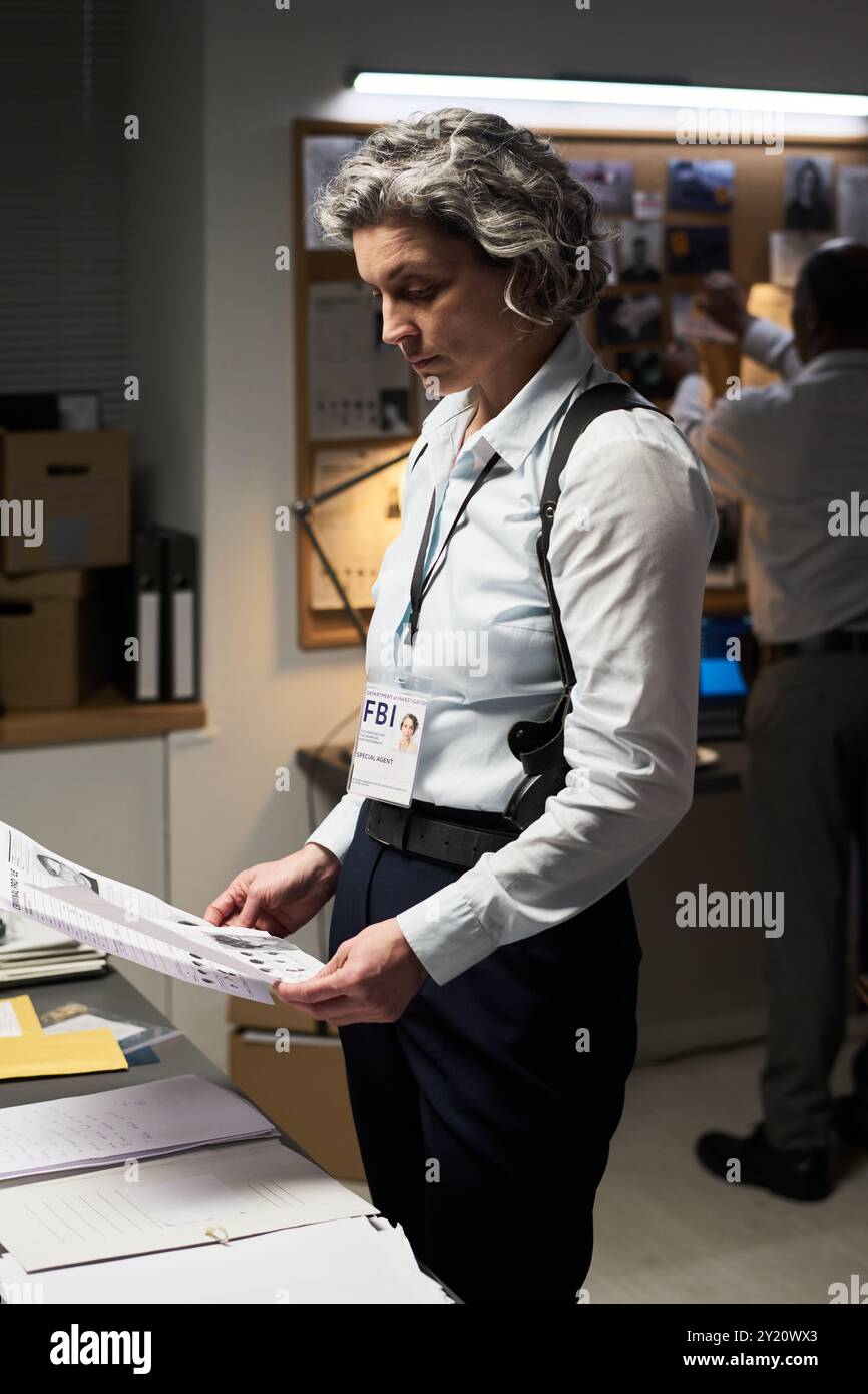 Analyzing crime scene evidence in modern office setting, person in white shirt wearing FBI badge examining documents under focused lighting creating a professional atmosphere Stock Photo