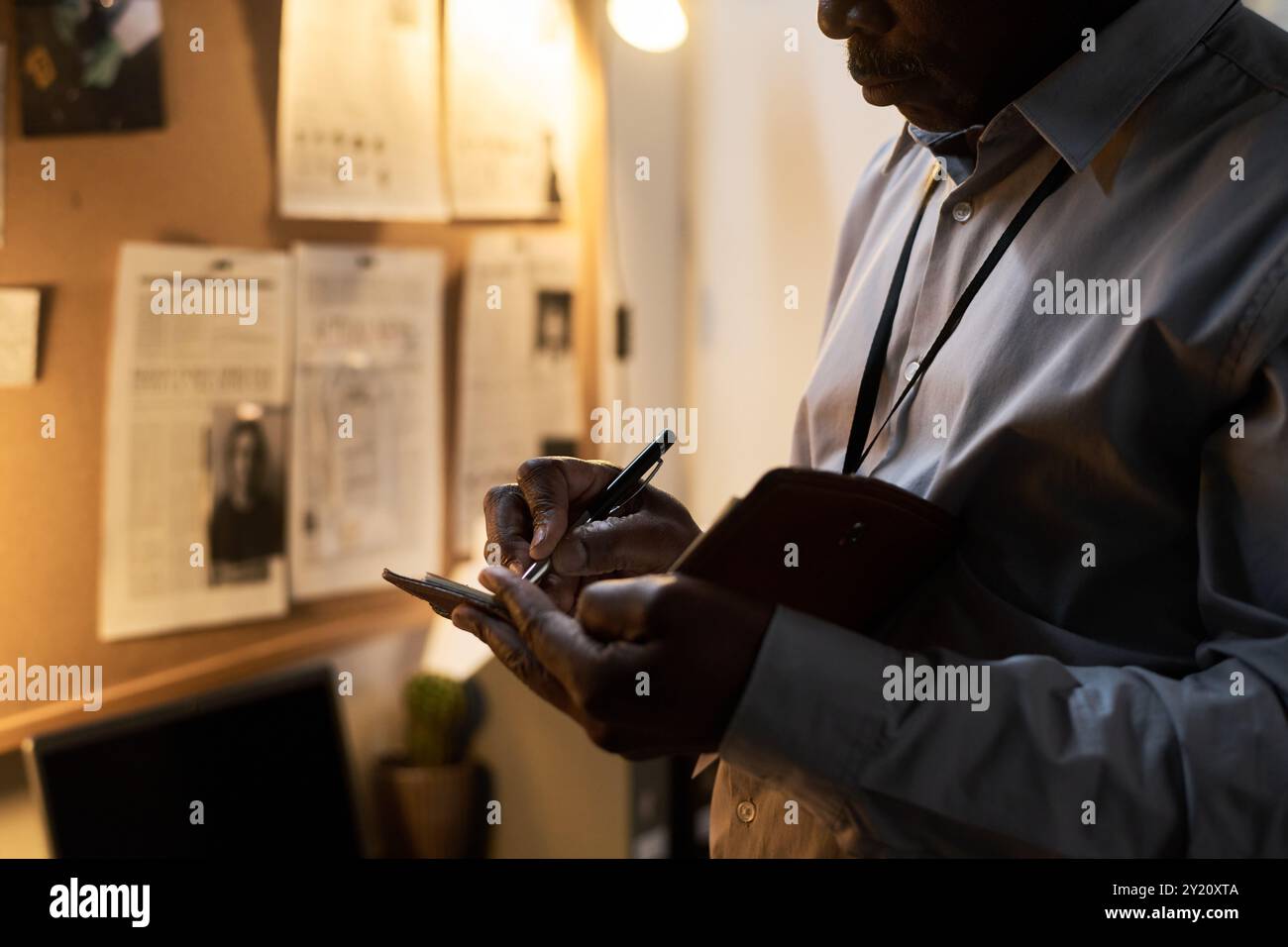 Man taking notes in office while reviewing documents on wall with focused expression and office lit warmly by desk lamp Stock Photo