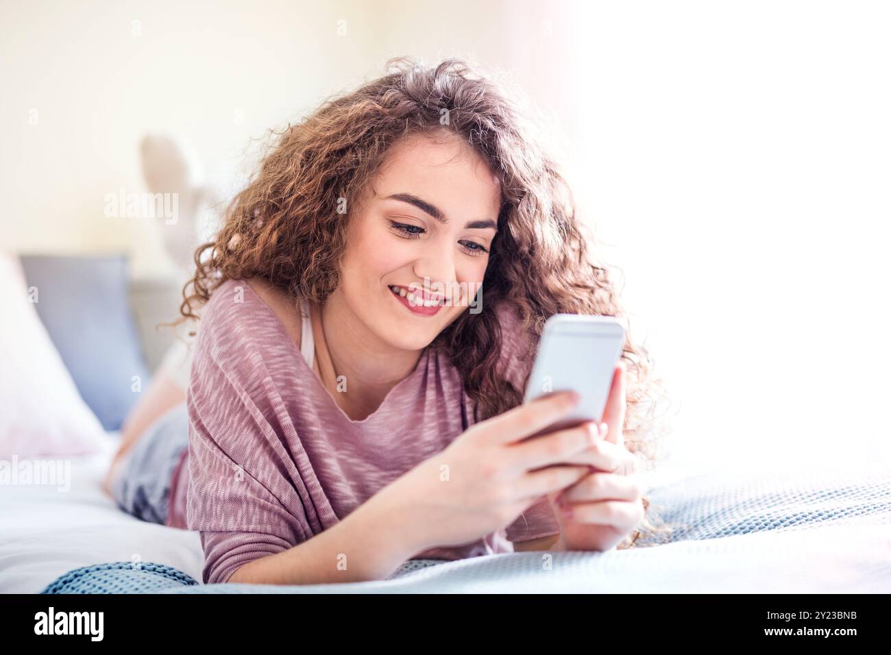 Beautiful young woman lying on her stomach on bed, scrolling on smartphone, smiling. Stock Photo