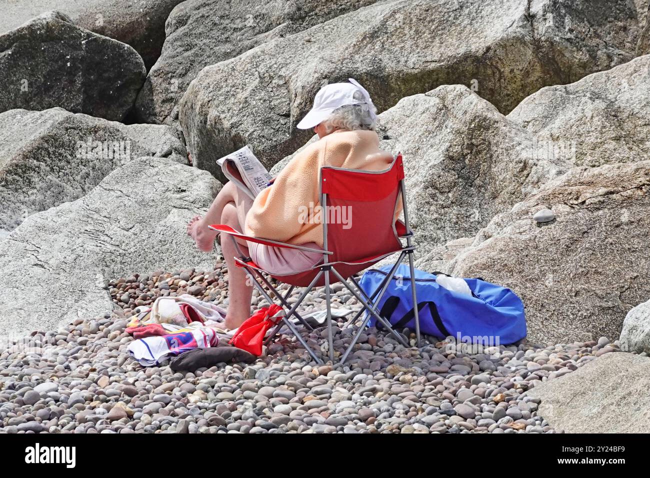 Senior citizen woman sitting alone beside breakwater rocks on pebble beach sunny day wrapped up against cool June sea breeze Sidmouth Devon England UK Stock Photo