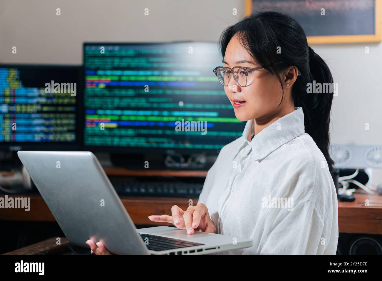 A young woman with glasses working on a laptop in a tech environment. She appears focused and engaged, with computer screens displaying code in the ba Stock Photo