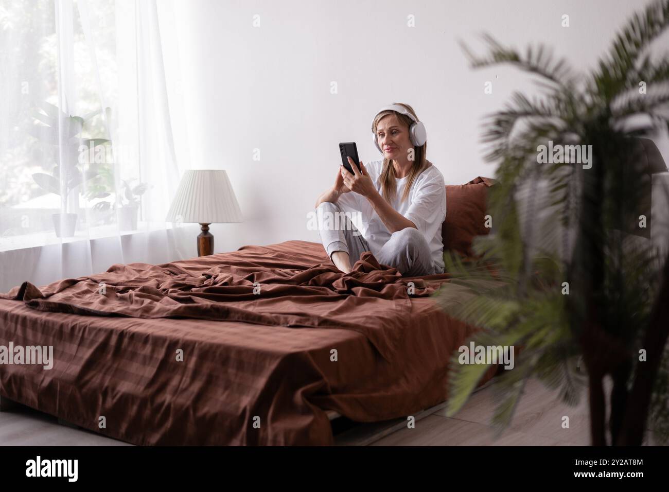 Cheerful older woman in headphones looks at smartphone, lies on brown bed listen favorite music. Relax, rest and lifestyle Stock Photo