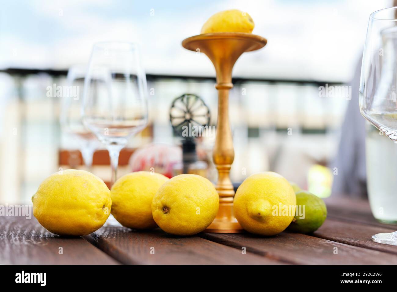 Pile of many bright ripe italian yellow lemons arranged on rustic wooden table wine glasses and decorative stand at rural countryside terrace patio Stock Photo