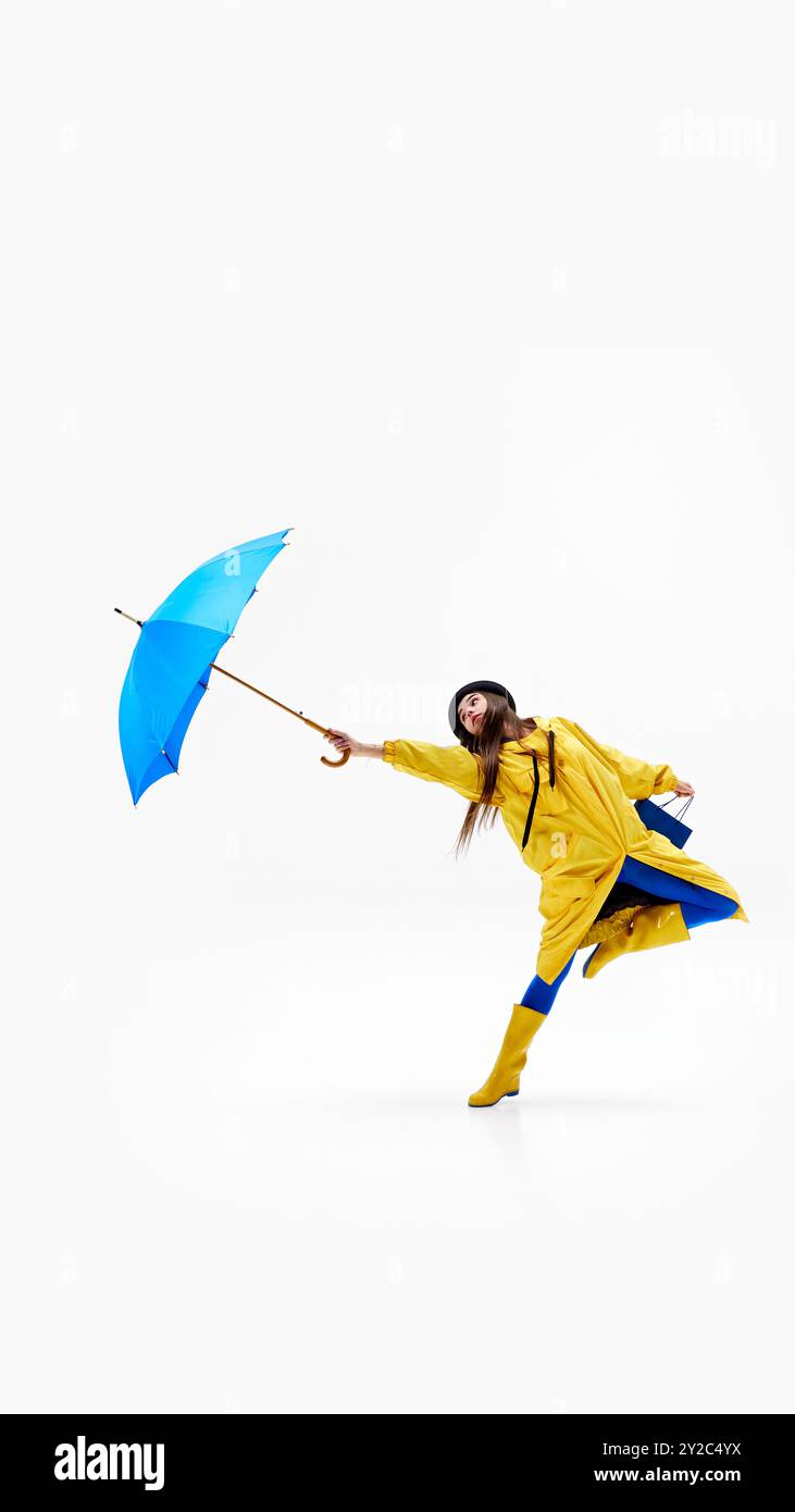 Cheerful woman in vibrant raincoat leaps with blue shopping bag and umbrella, embracing rainy weather against white background. Stock Photo