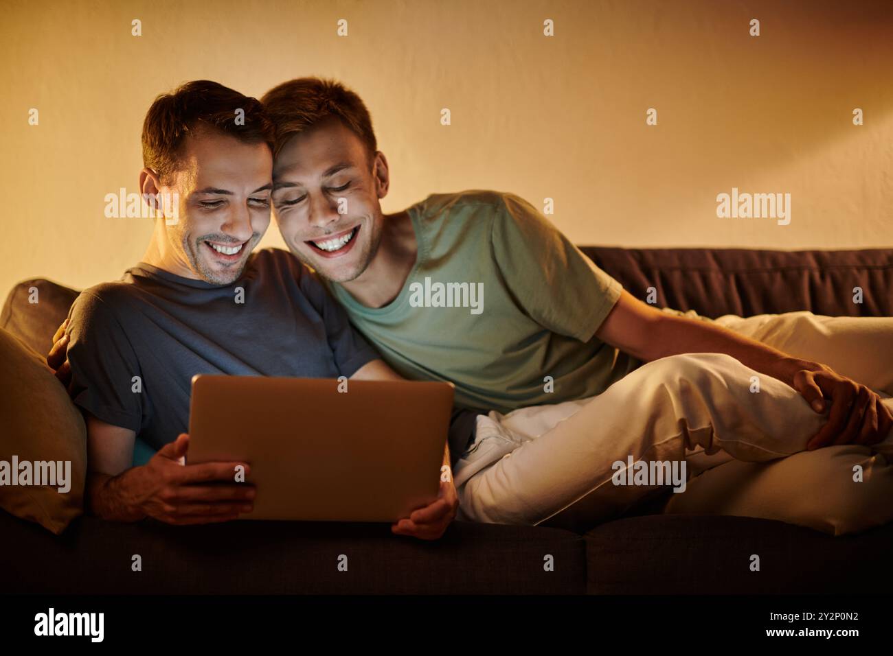 Two men share laughter and joy while using a laptop, embracing on the couch. Stock Photo