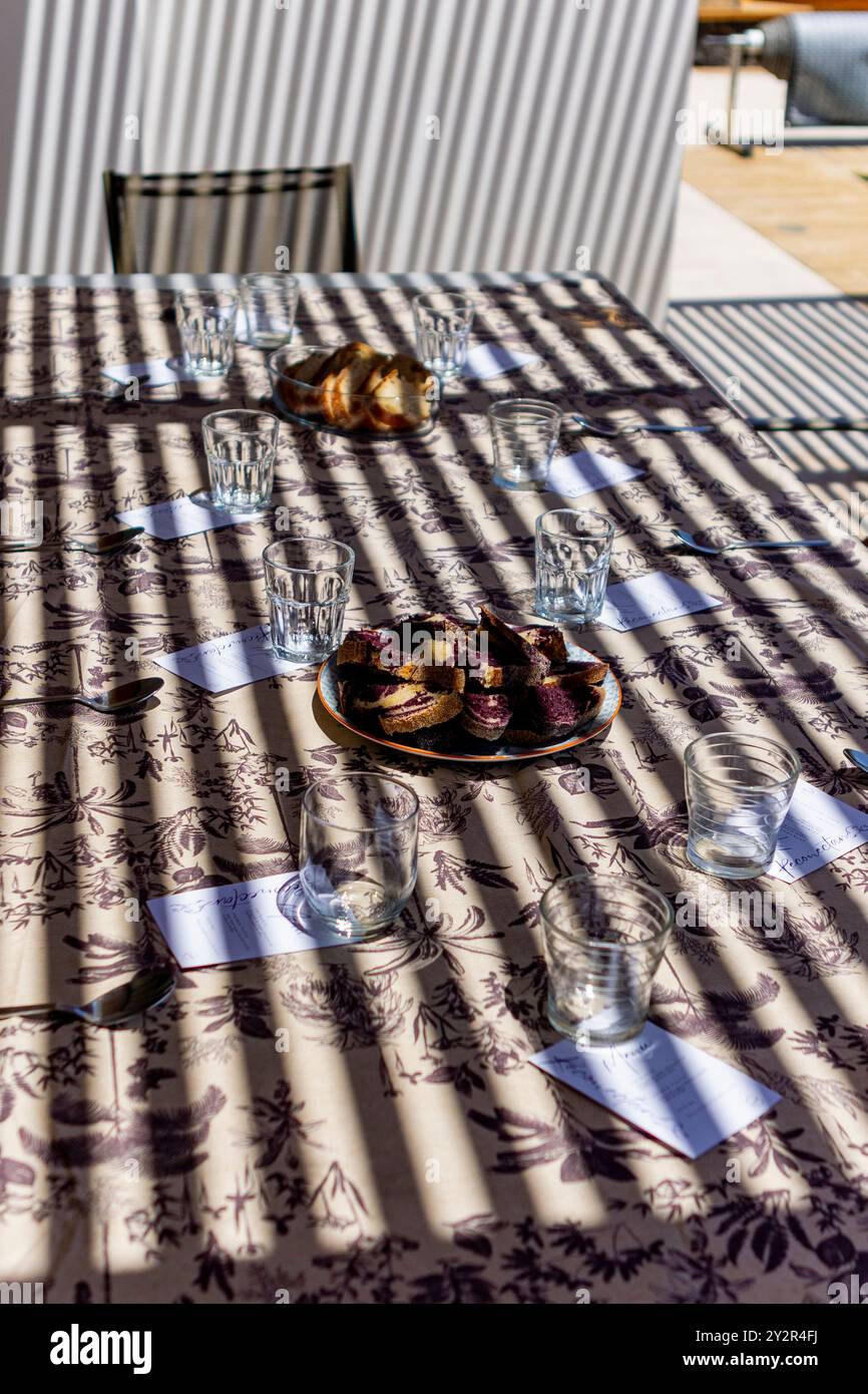 An outdoor dining table is bathed in sunlight, casting striking shadows across a patterned tablecloth. Slices of bread and beautifully-prepared food a Stock Photo