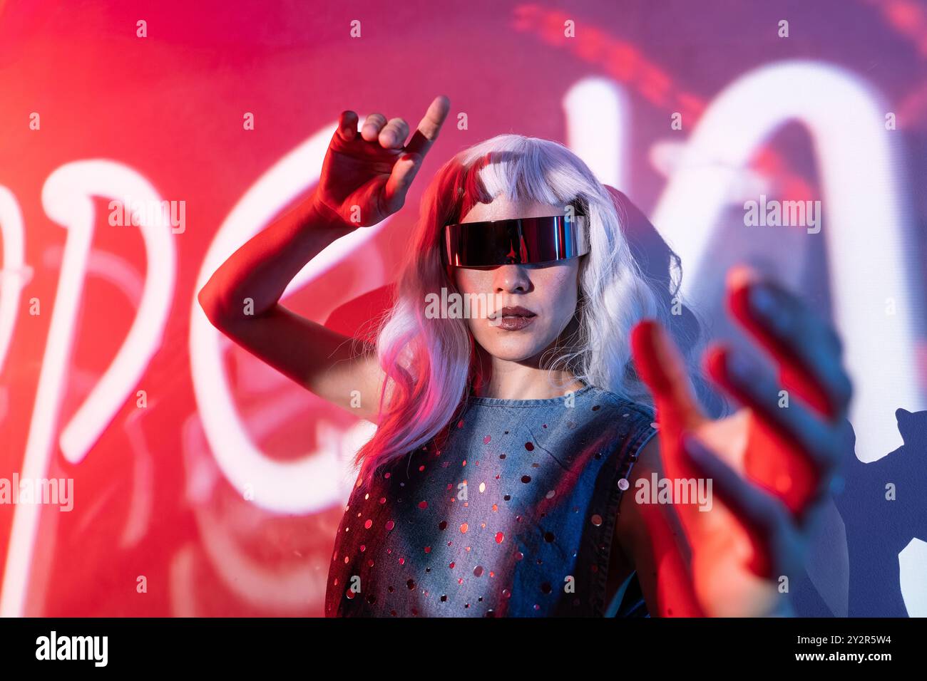 A young woman with silver hair and futuristic glasses stands against a vibrant red backdrop, actively interacting with a cutting-edge digital interfac Stock Photo