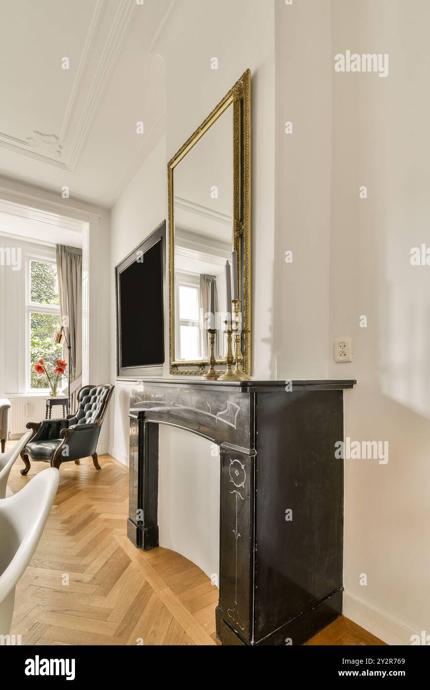 A refined living room featuring a black antique fireplace adorned with a large golden frame mirror and a cozy seating area by the window. Stock Photo