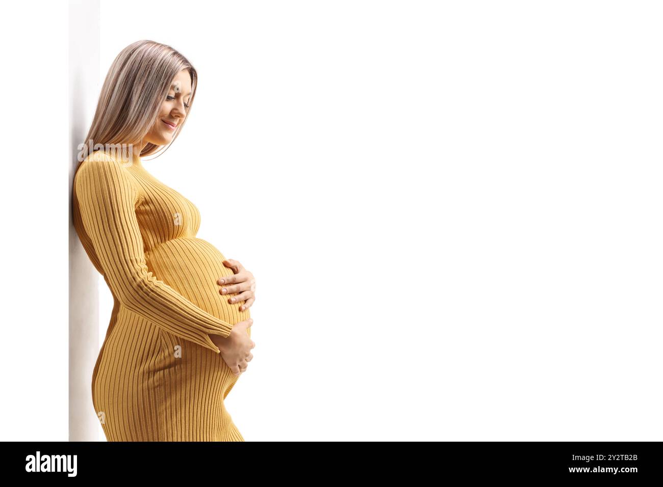 Beautiful pregnant woman in a yellow dress leaning on a wall and holding her belly isolated on white background Stock Photo