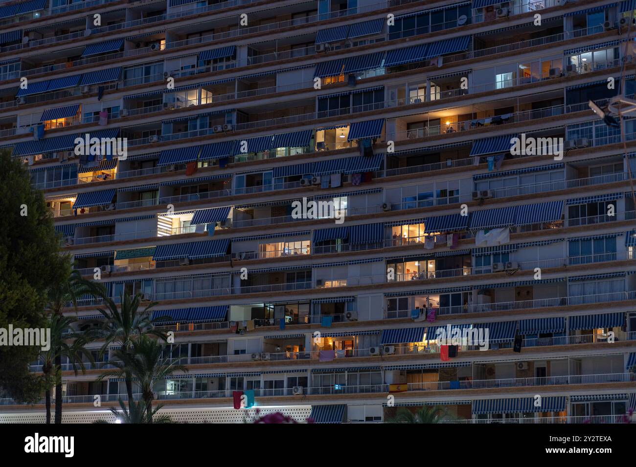 A residential building in Alicante, Spain with blue striped awnings and illuminated windows during nighttime, creating a peaceful urban scene. Stock Photo