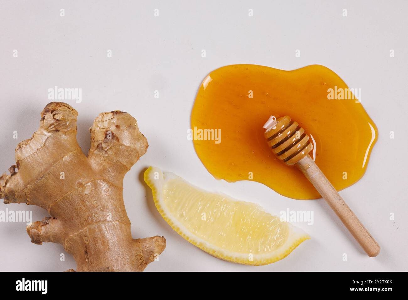 A flat lay of fresh ginger root, a slice of lemon, and honey drizzled on a white surface. A small wooden honey dipper rests in the honey, creating a n Stock Photo