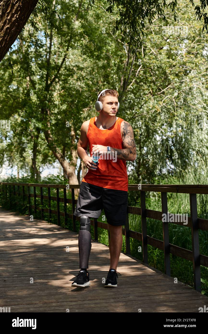 A young man with tattoos and prosthetic leg stands on bridge, with water bottle, listening music surrounded by park greenery. Stock Photo