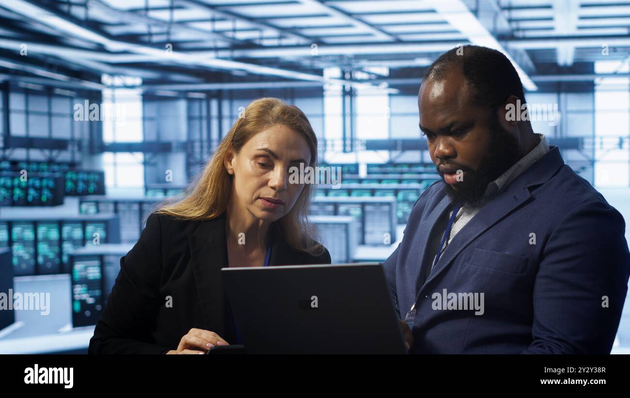 Teamworking colleagues in server farm using laptop to patch network issues affecting transfer rates. Coworkers in data center doing brainstorming, fixing errors affecting supercomputers, camera A Stock Photo