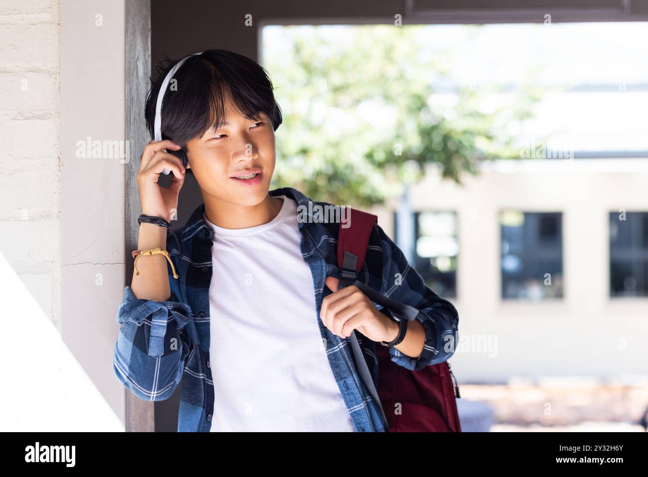 Listening to music with headphones, asian teenage boy leaning against wall at school Stock Photo