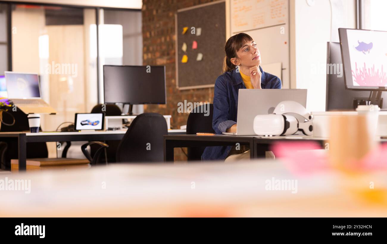 Working in office, woman using laptop and thinking, surrounded by monitors Stock Photo