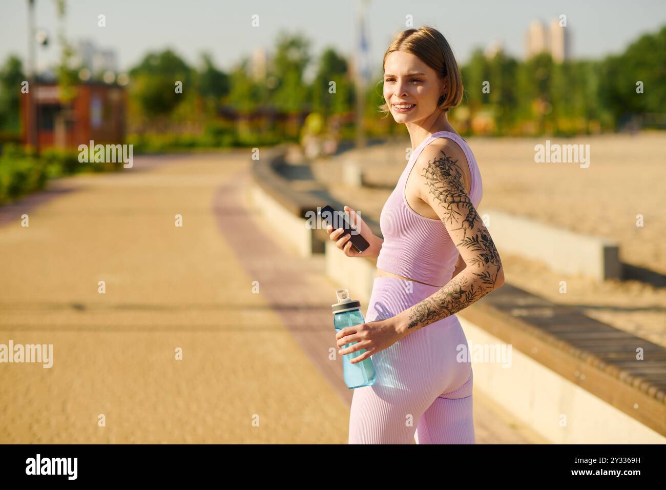 A cheerful young woman walks through a vibrant park with a water bottle in hand. Stock Photo