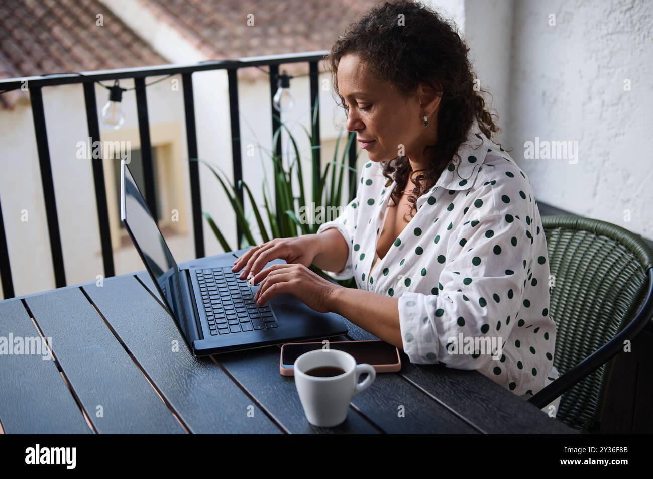 A woman focuses on her laptop while working remotely on a cozy balcony ...