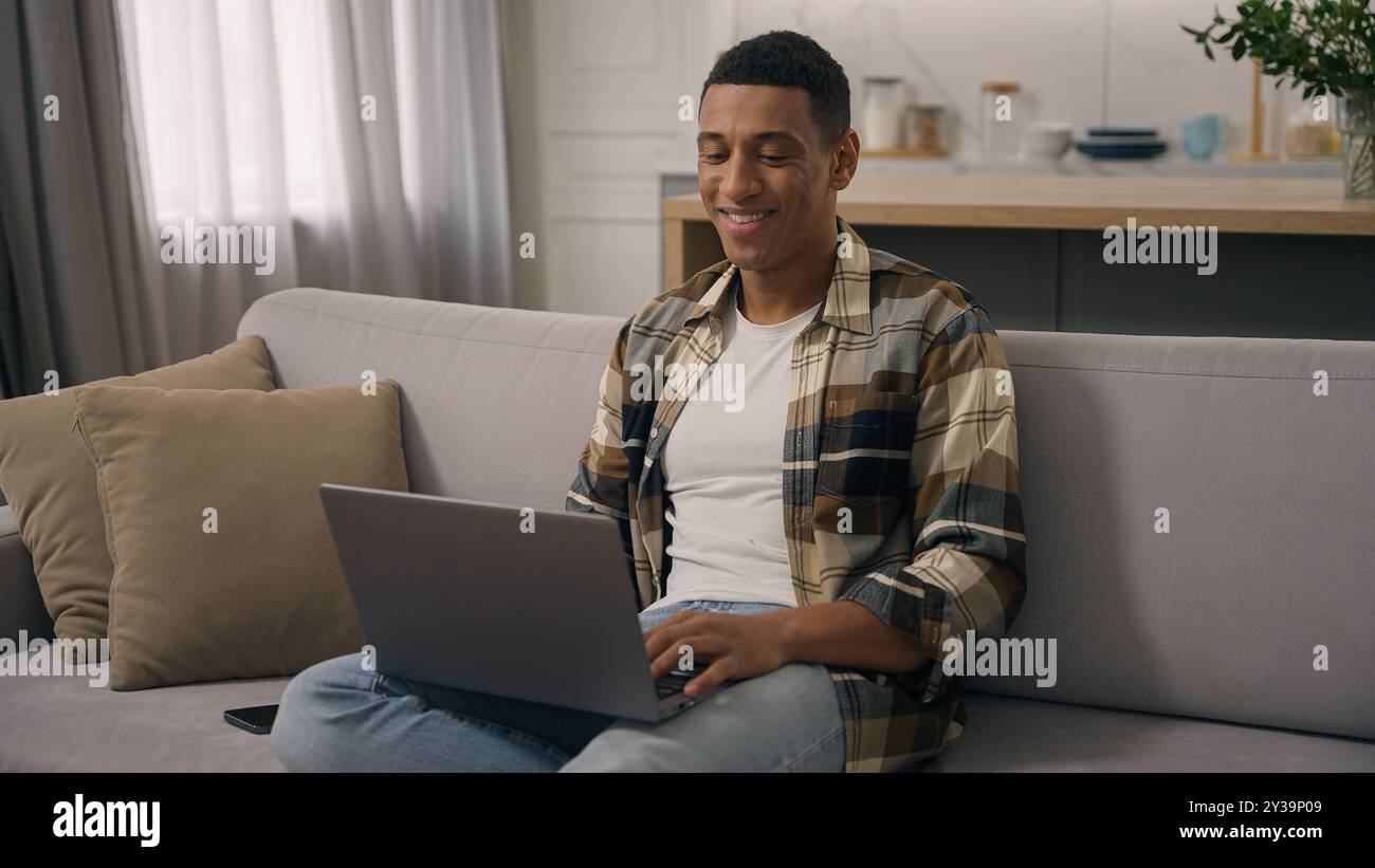 Happy smiling African American ethnic man working at home distant online work studying at sofa guy laptop using computer at couch male using pc Stock Photo