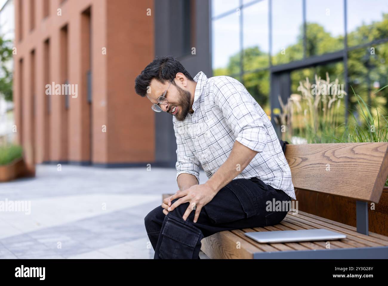 Winced Image A young Indian man sits outside a building on a bench with his hands on ...