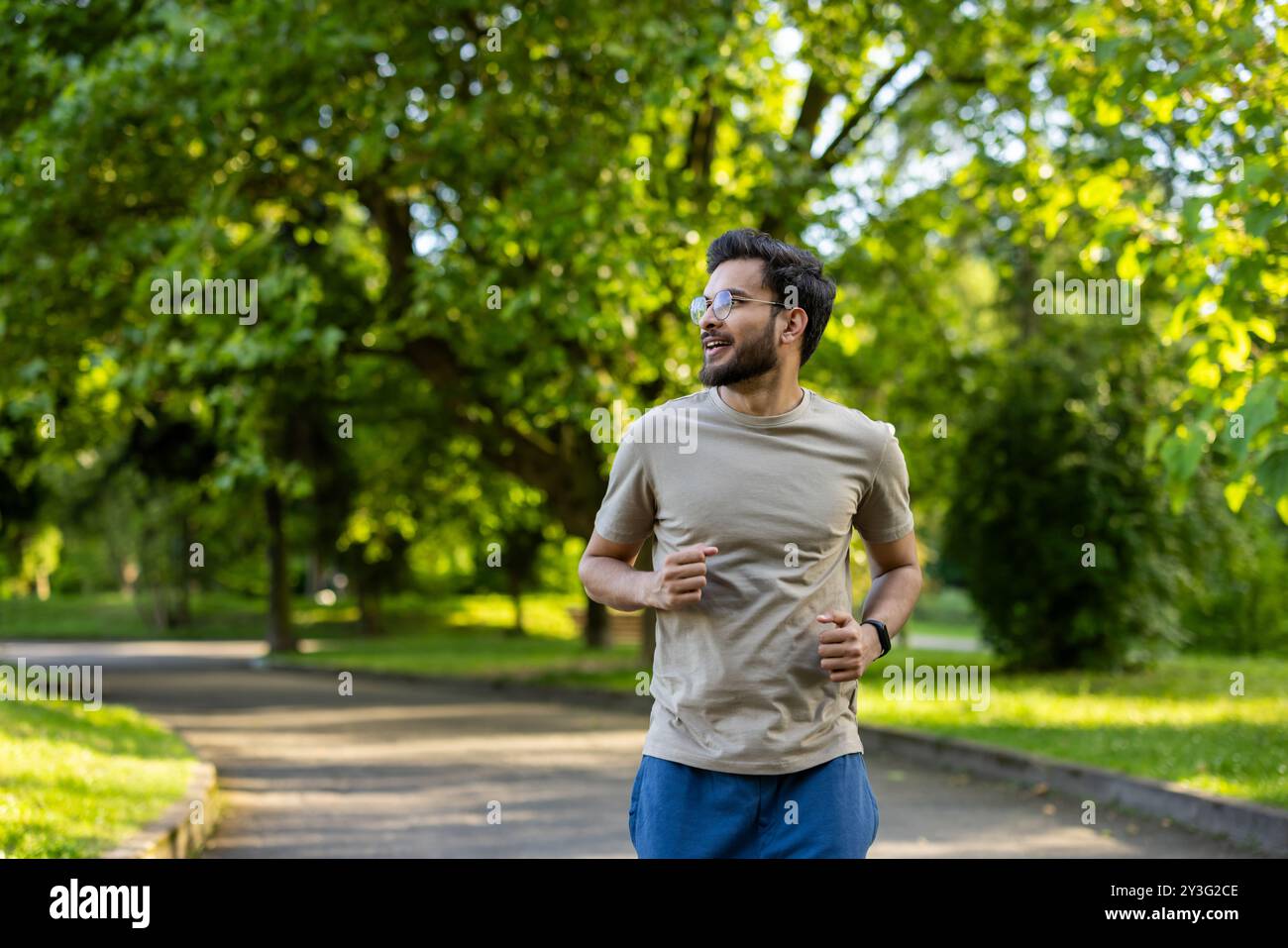 Young sportsman running through peaceful park, appearing cheerful and energized. Casual wear indicates enjoyment of outdoor exercise. Morning sun filters through green leaves Stock Photo