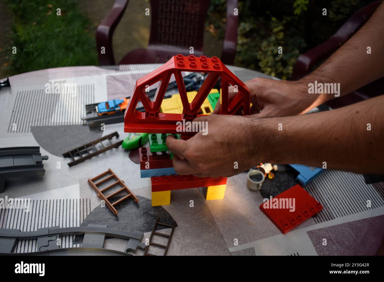 A man assembling toy  bricks on a garden table with colourful tablecloth in a garden. Stock Photo