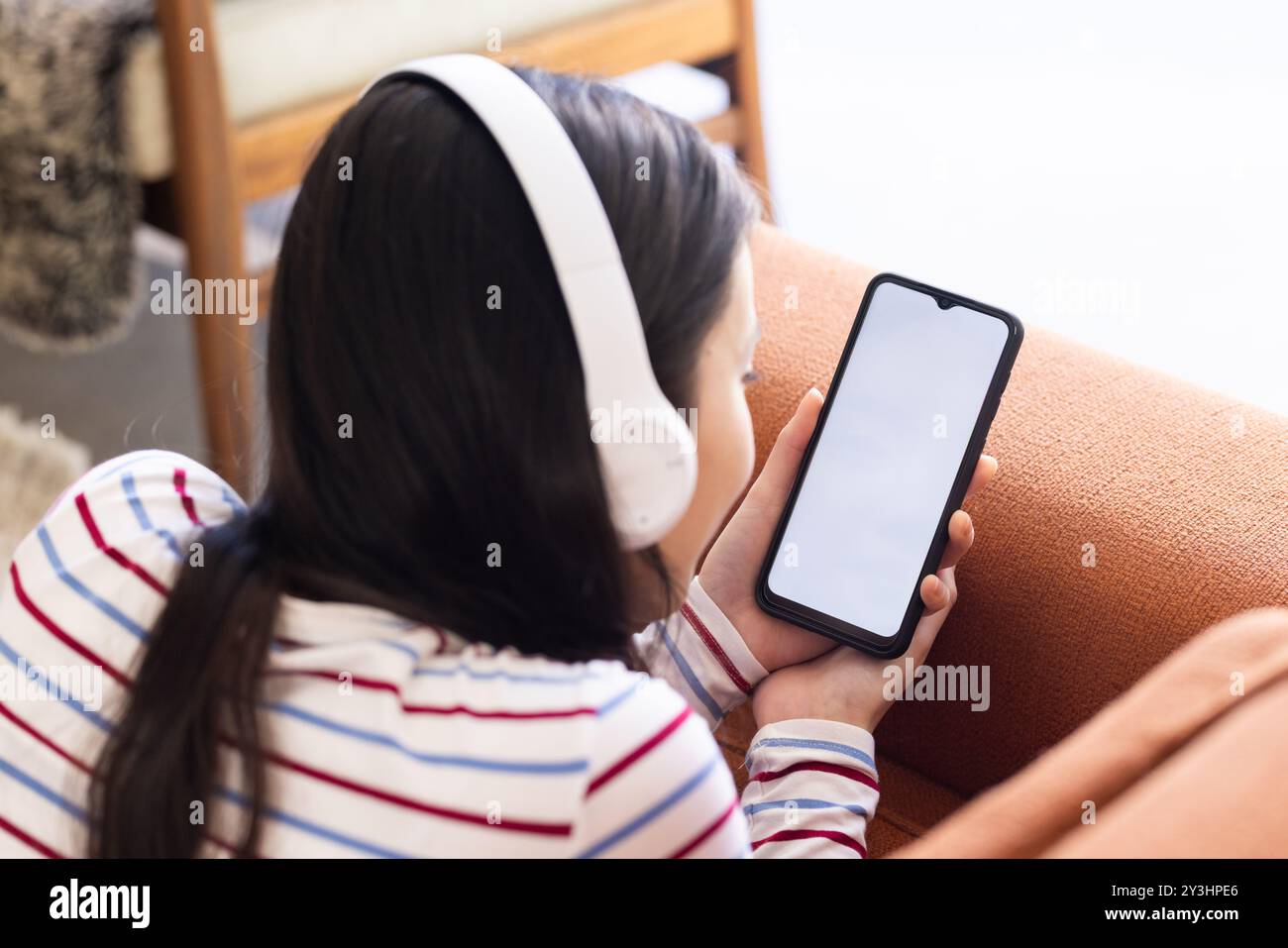 At home, wearing headphones, young asian woman using smartphone while sitting on couch, copy space Stock Photo