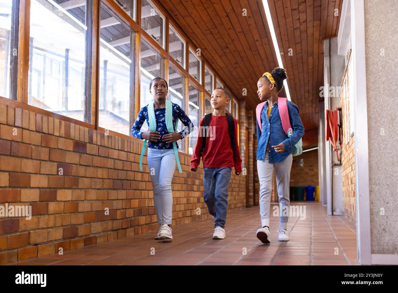 Walking in school hallway, two multiracial girls and boy with backpacks talking and smiling Stock Photo