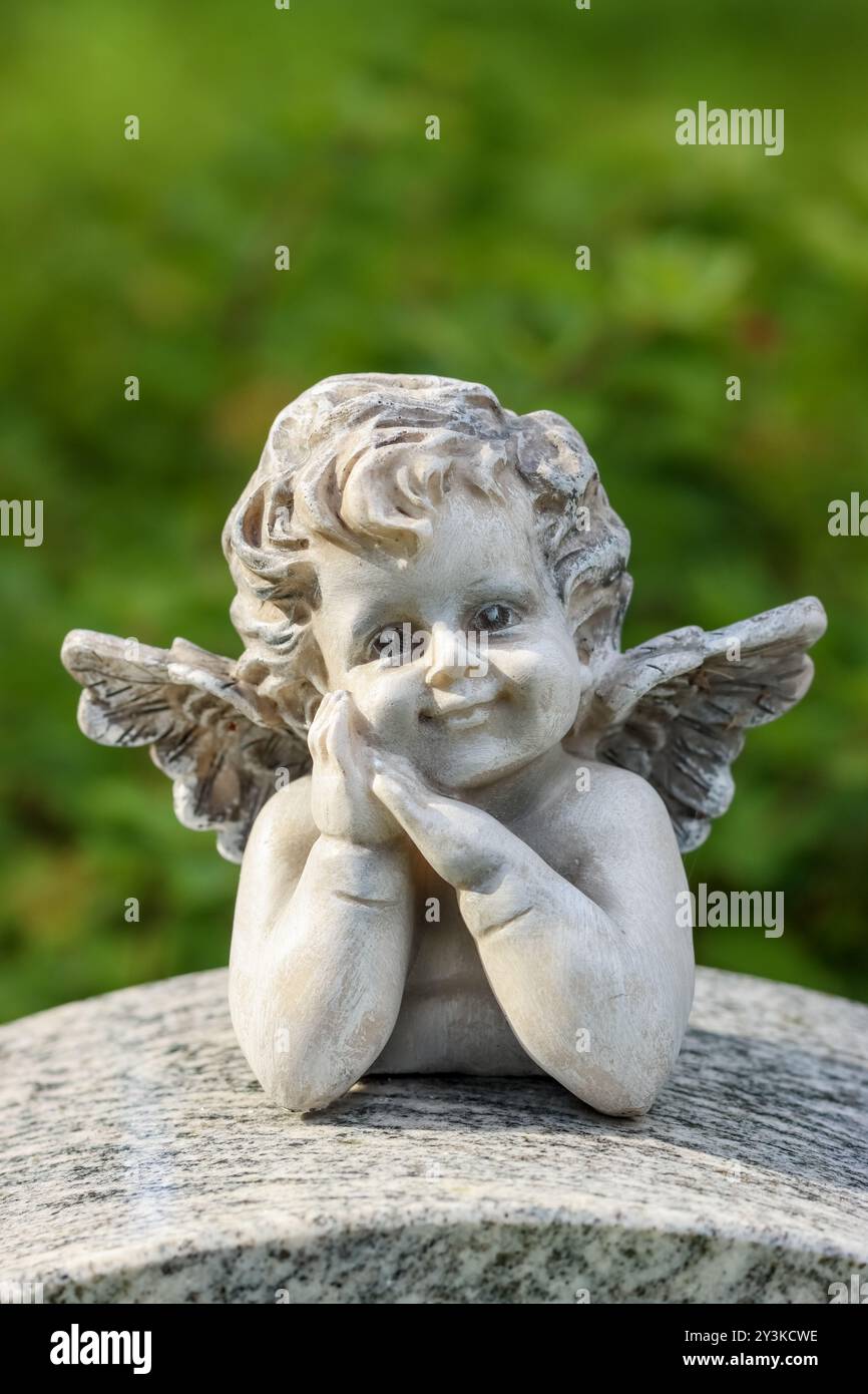 A small angel statue as a grave decoration in a cemetery. The picture has an empty space for text. The statue has a green summer background. Stock Photo