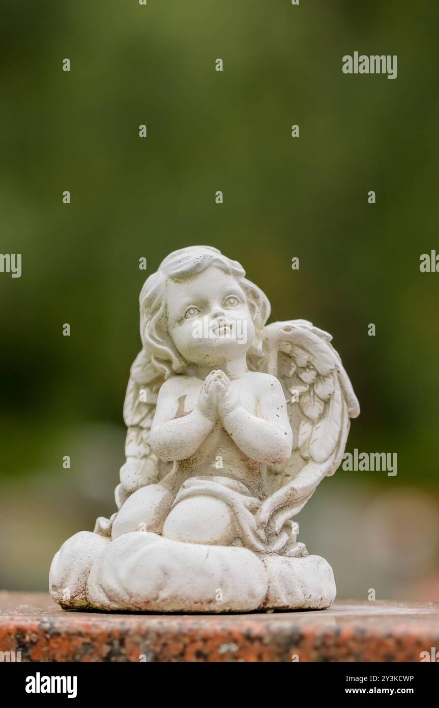 Small white angel statue as a grave decoration in a cemetery. The picture has an empty space for text. The statue has a green summer background. Stock Photo