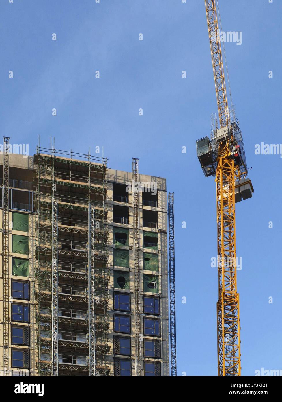 A tall yellow construction crane working on a large hight rise concrete building with scaffolding against a blue sky Stock Photo