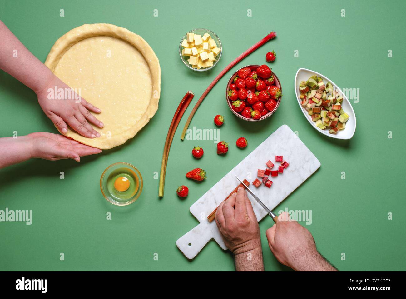 Above view with young people hands making a sweet pie with organic ingredients. Strawberries and rhubarb cake making of, on a green table, top view Stock Photo