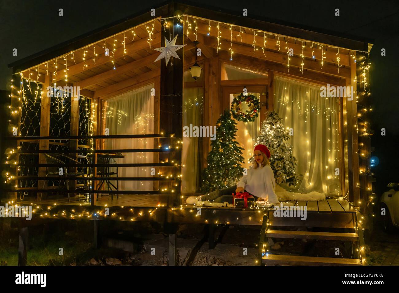 Christmas Lights Cabin Woman - A cozy cabin decorated with Christmas lights, a woman in a Santa hat sitting on the porch steps at night. Stock Photo