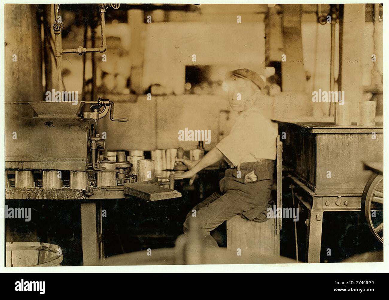 Name: Dangerous Business. Boy working at canning machine with open gearing. Many suck machines and boys too. J.S. Farren & Co. Witness--J. W. Magruder. July 7, 1909. Location: Baltimore, Maryland. Stock Photo