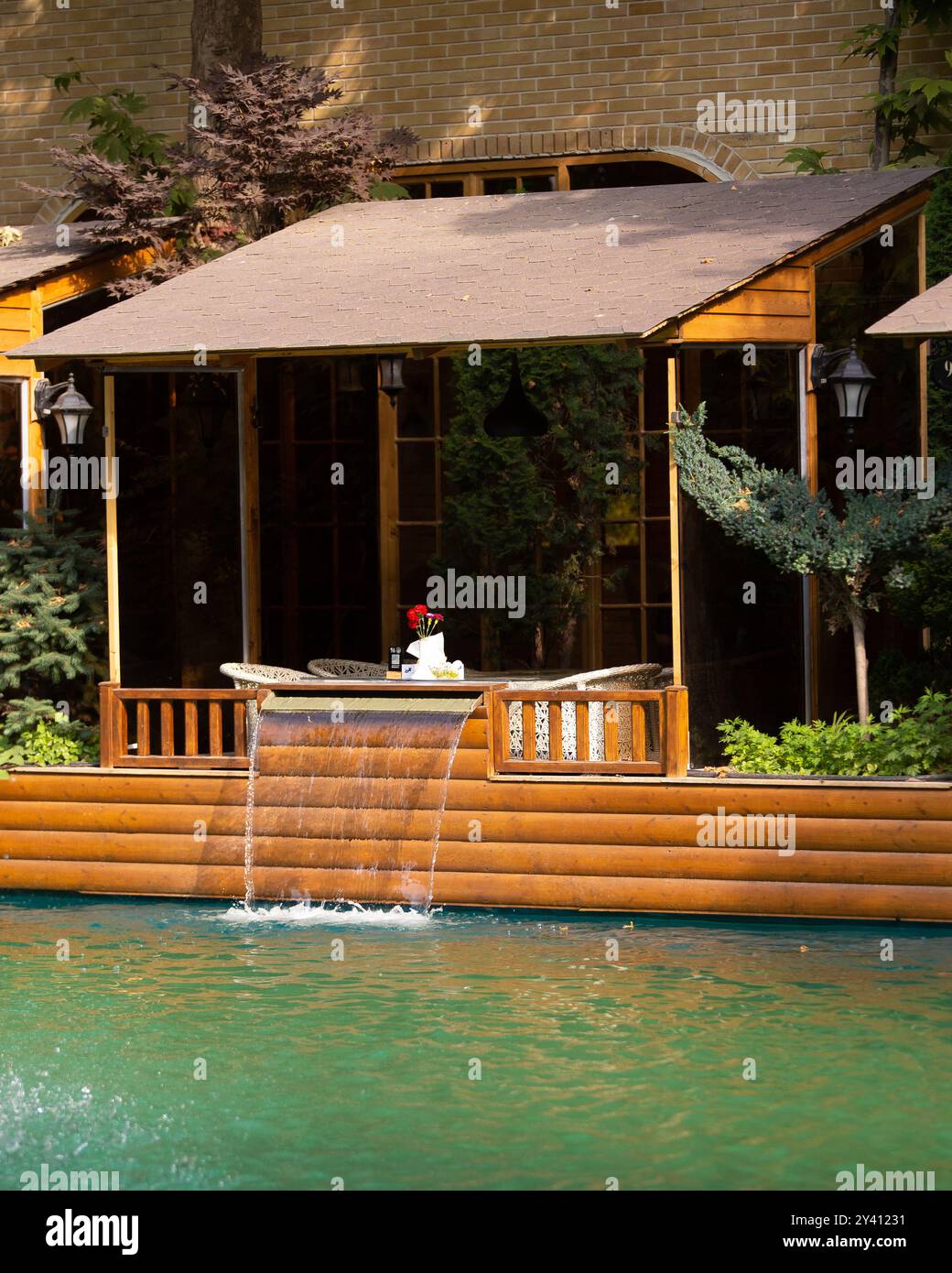 A peaceful waterside setting with a wooden cabin and cascading waterfall into a turquoise pool, ideal for relaxation. Stock Photo