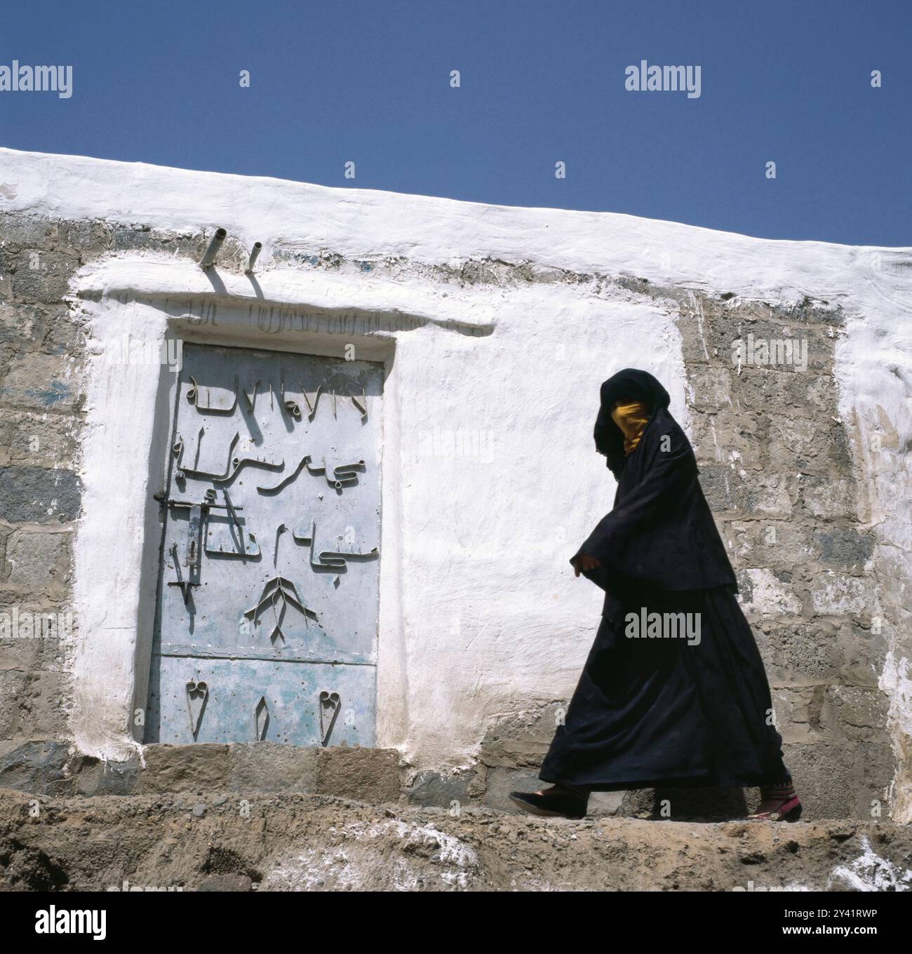 Yemeni woman in traditional veil walking alongside a wall in Sanaa with a door with decorated ornaments Stock Photo