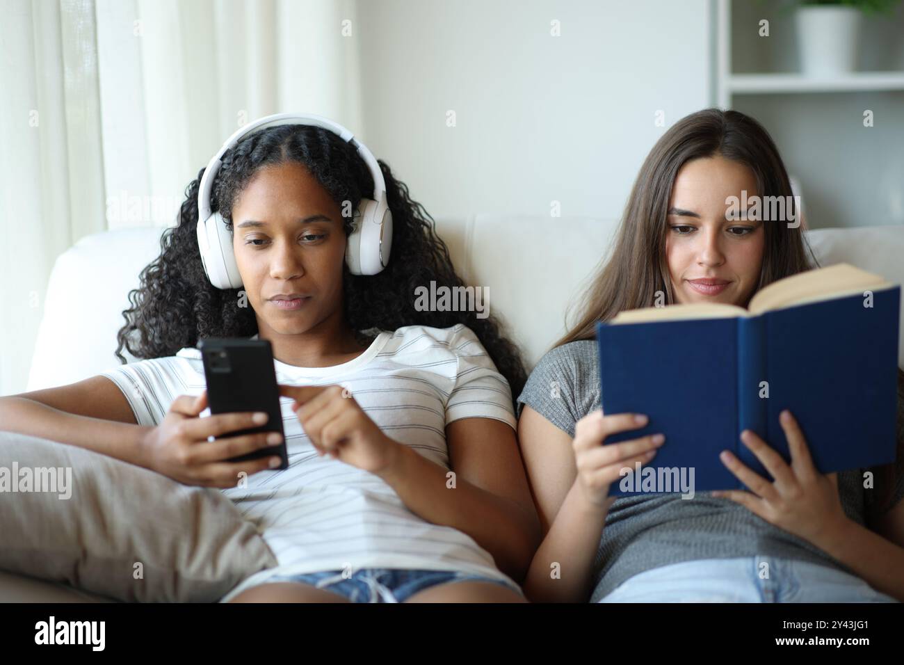 Front view portrait of two interracial roommates one is listening music and the other reads a paper book Stock Photo