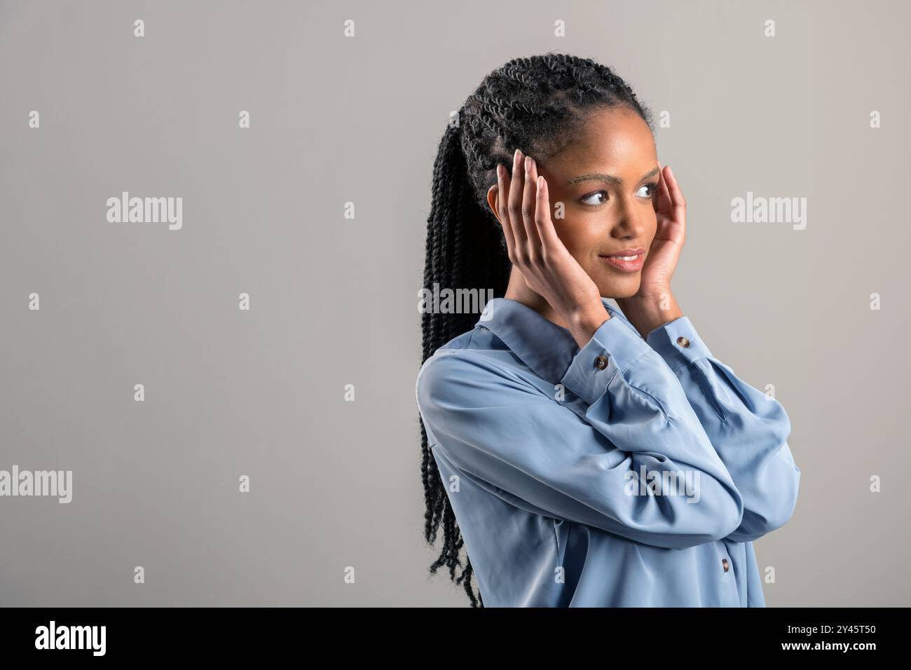 Smiling young African American female with long braided hair looking away while touching cheeks against gray wall Stock Photo