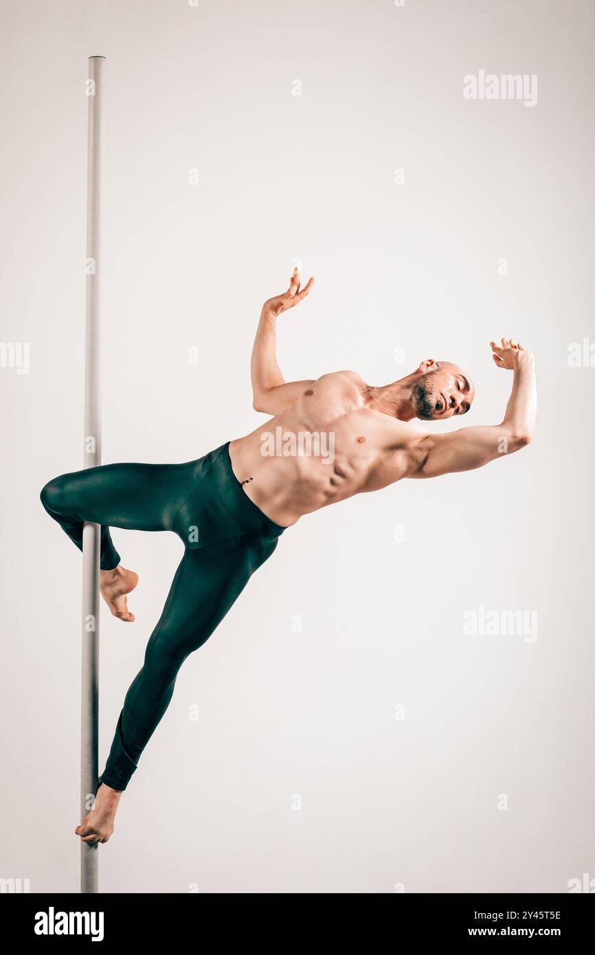 Full body of muscular male pole dancer gracefully holding the cupid pose showcasing strength and flexibility against colored background Stock Photo