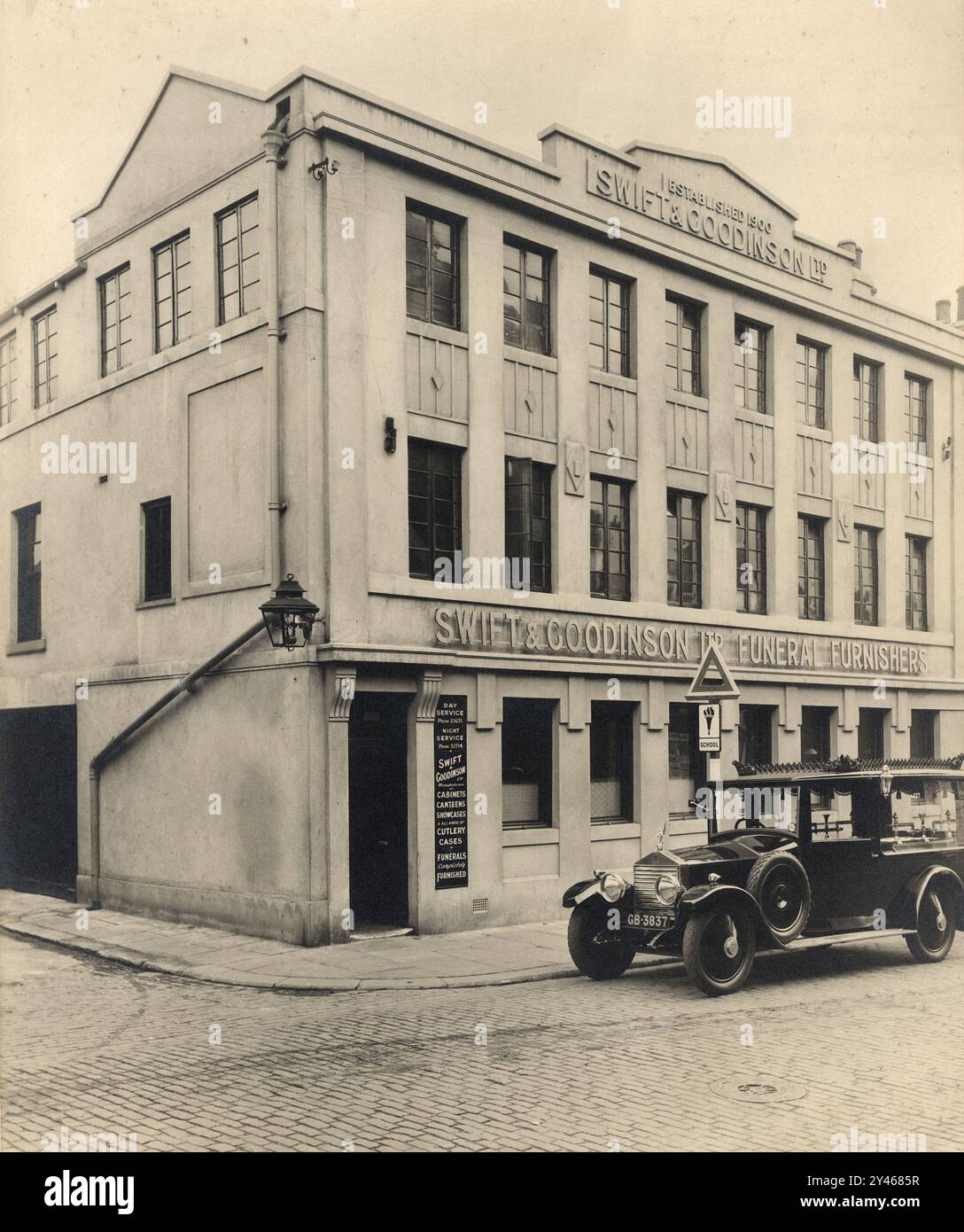 Swift Goodinson Funeral Furnishers, photograph of large premises, 1920s.  Black Rolls Royce hearse outside.   Firm established 1900 in Sheffield, england.  As well as all funeral needs, they made cutlery cabinets and cases for the Sheffield trade.  Gas street lamp. Cobbled road. Vintage road sign for School on pavement. Stock Photo