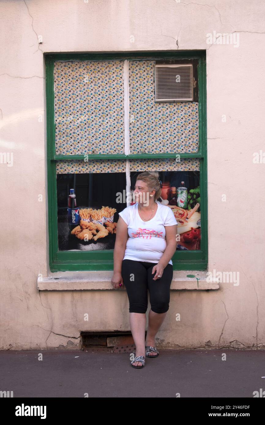 Smoking cigarettes outside, a middle aged woman smoked sitting on window  sill outside of a grocery shop and general store. In the window there are  large poster photographs of the food thats
