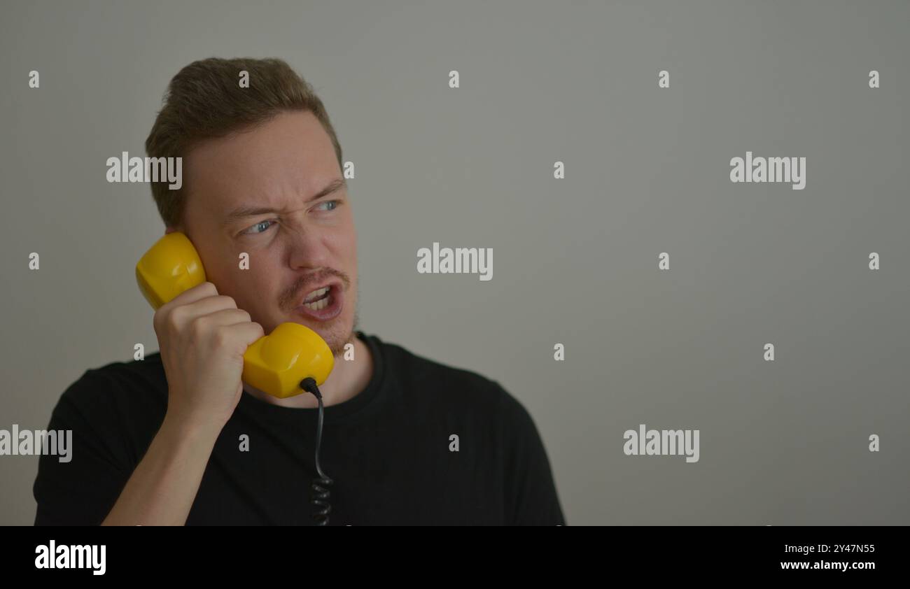 A man talking and shouting on the phone with a yellow retro telephone receiver. Emotion expression of anger concept. Emotional reaction from the conve Stock Photo