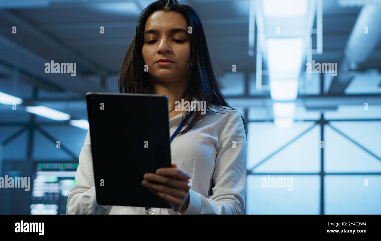 Software developer walking in data center, using tablet to design, implement and manage gear, ensuring optimal performance and reliability. IT worker in server farm coding on device, camera A Stock Photo