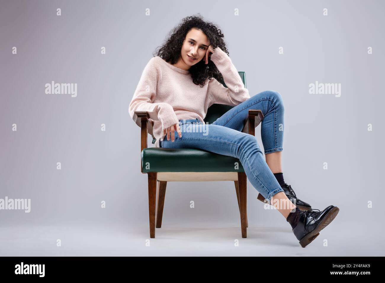 Young woman with curly hair is posing sitting on a vintage armchair with her legs crossed, her left hand resting on the armrest and the right one on h Stock Photo